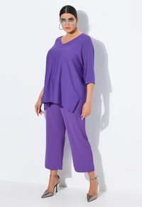 Haut violet surdimensionné avec un col en V, associé à un pantalon violet crop et des chaussures à talons hauts argentées. Lunettes de soleil transparentes et grandes boucles d'oreilles.