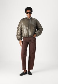 Olivenfarbene Puffjacke mit glänzender, gesteppten Textur und gerippten Bündchen, kombiniert mit braunen, hochtaillierten Jeans und schwarzen Slippern.