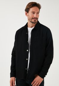 Camisa de mezclilla negra con botones en la parte delantera, un bolsillo en el pecho y mangas largas. Textura suave, ajuste entallado y botones de presión de metal.