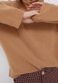 Personne portant un pull à col roulé couleur chameau avec des poignets côtelés, les bras croisés sur un pantalon à motifs bordeaux et beige avec un bouton doré.