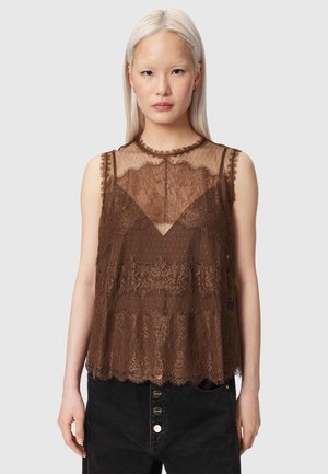 Vrouw met stijl blond haar draagt een bruine kanten mouwloze top over een bruine bralette, gecombineerd met zwarte high-waist jeans met zichtbare knopen.
