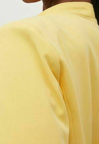 Keltainen kangas, jossa on pehmeä rakenne, pyöreä pääntie ja hieman puffatut olkapäät. Materiaali näyttää kevyeltä ja sileältä.