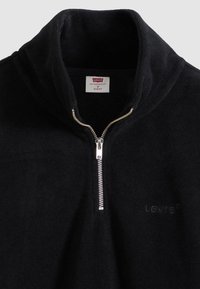 Μαύρο φλις μπλουζάκι με φερμουάρ στο ένα τέταρτο και ψηλό γιακά. Διαθέτει ασημί φερμουάρ και διακριτικό ανάγλ υφο λογότυπο. Μαλακή υφή και σχεδόν εφαρμοστό σχέδιο.