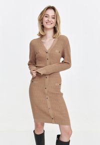 Dzianinowa sukienka z długim rękawem w jasnobrązowym kolorze, z dekoltem w kształcie litery V, zapinana na guziki, z teksturowanym wzorem oraz dwiema kieszeniami z przodu z czarnymi akcentami.