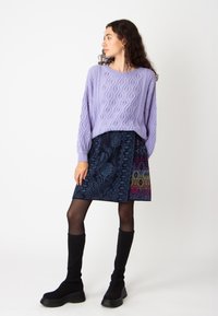 Tricot violet en laine avec un motif en cable, associé à une jupe marine à motifs comportant des designs floraux et géométriques, complétée par des bottes noires montant au genou.