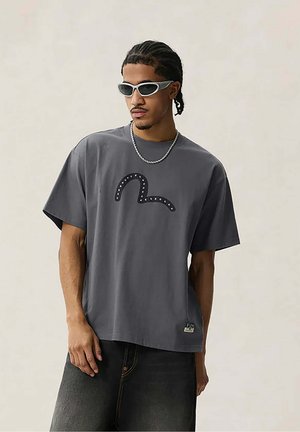 Uomo con capelli intrecciati che indossa una t-shirt grigia con grafica, una catena d’argento, occhiali da sole scuri e jeans neri su uno sfondo semplice.