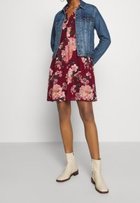 Un vestido floral en burdeos con flores rosas, combinado con una chaqueta de mezclilla azul y botas de tobillo color crema, mostrando un estilo casual de verano.