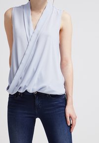 Blusa senza maniche azzurro chiaro con design drappeggiato a incrocio. Realizzata in tessuto morbido, con scollatura a V e una texture delicata.