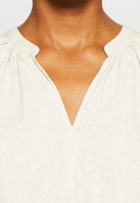 Blusa color blanco roto con escote en V, detalles fruncidos en los hombros y un tejido ligero y texturizado. Diseño simple y minimalista.