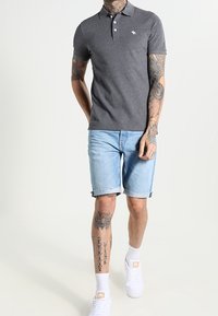Muž v tmavě šedém polo tričku, světle modrých denimu šortkách, bílých ponožkách a bílých teniskách, s tetováním na rukou a nohách, walking proti jednolitému pozadí.