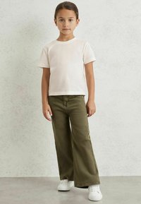 T-shirt bianco a maniche corte, jeans larghi verde oliva e sneakers bianche indossati da un bambino. Sfondo semplice con una texture sottile.