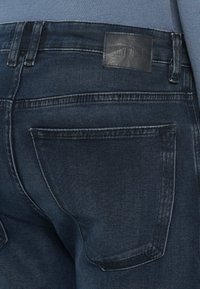 Pantalones vaqueros de mezclilla azul oscuro con el bolsillo trasero con costura visible y una etiqueta de marca de cuero negra en la cintura.