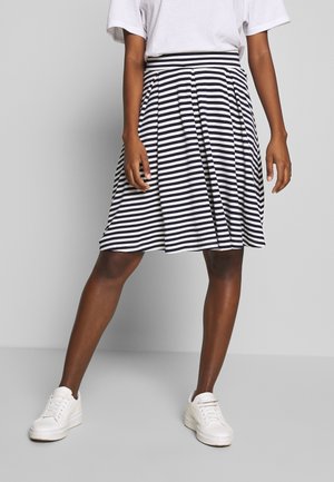 Woman wearing a black and white horizontal striped knee-length skirt, white t-shirt, and white sneakers standing with one leg slightly forward.