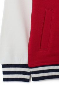 Maglione lavorato a maglia rosso e bianco con polsini a coste blu navale e bianchi a righe orizzontali, dotato di una tasca laterale nella parte rossa.