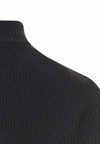 Donkere geribde trui met een hoge, gestructureerde halslijn. Stof lijkt zacht met een lichte glans. Close-up toont textuur en stikseldetails.