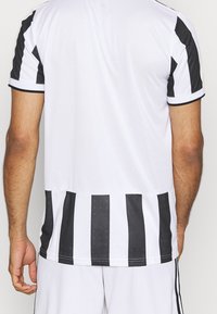 T-shirt de sport blanche avec des rayures verticales noires sur les manches et le bas du dos. Fabriquée en tissu léger, elle présente un col rond et des manches courtes.