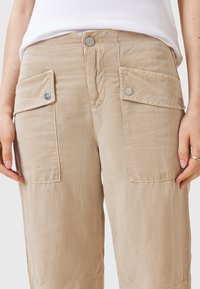 Beige cargo kalhoty vyrobené z lehkého materiálu, s dvěma velkými bočními kapsami, stříbrnými patentkami a volným střihem.