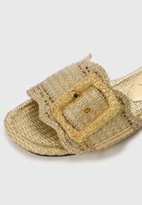 Gold-toned slide sandal made of woven material with a textured finish, featuring a large, decorative buckle and scalloped edges.