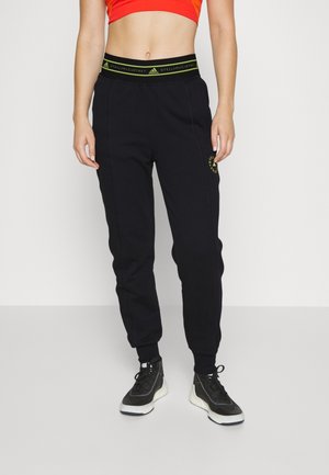 Personne portant un pantalon de jogging noir Adidas by Stella McCartney, des baskets noires et un top de sport court rouge, debout sur un sol gris.