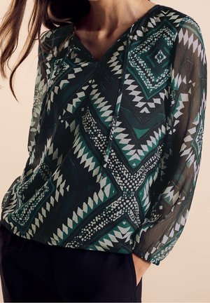 Vrouw die een doorschijnende lange mouwen blouse draagt met groene, zwarte en witte geometrische patronen en een zwarte broek, hand in zak.