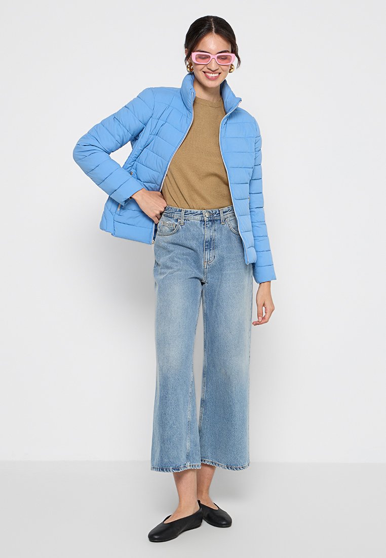 Himmelblaues Puffermantel mit horizontalen Nähten, goldbrauner Pullover, weite helle Jeans und schwarze Ballerinas. Rosa übergroße Sonnenbrille.