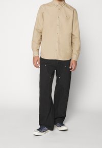 Camisa de manga larga color arena con una textura suave, combinada con pantalones negros de corte holgado con detalles dorados y zapatillas blancas con un diseño azul.