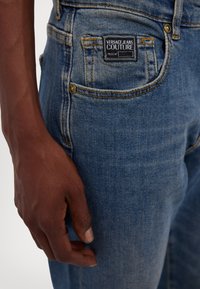 Világoskék farmernadrág, derékban szűkített fazonnal, hátul zsebében címkével, kontrasztos varrással és fémből készült gombos részlettel.