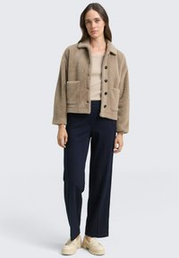 Beżowa pluszowa kurtka z kołnierzem, dwoma przednimi kieszeniami i czarnymi guzikami. Nosi się ją na jasnobeżowej bluzce w połączeniu z ciemnoniebieskimi spodniami.