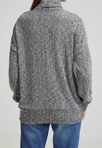 Grå och svart mönstrad tröja med hög krage, lös passform och långa ärmar, kombinerad med blå jeans. Texturerad stickad tyg.