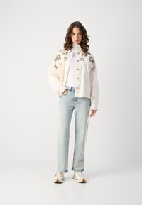 Kremfarget jakke med blomsterbroderi, knappelukking, sammen med lyseblå jeans og joggesko. Antrekket har en avslappet passform.
