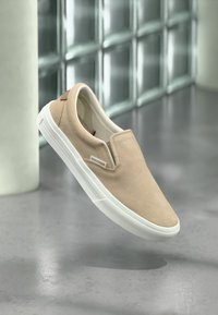 Μπεζ σουέτ παπούτσι slip-on με λευκή καουτσούκ σόλα, που διαθέτει μικρή ταμπέλα με λογότυπο και καφέ λεπτομέρεια στη φτέρνα. Ομαλή υφή σε όλη την επιφάνεια.