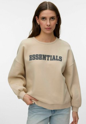 Ženska z dolgimi rjavimi lasmi, oblečena v bež pulover z napisom "ESSENTIALS" in svetlo modre kavbojke, stoji ob belem ozadju.