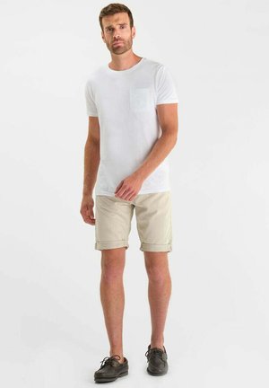 Uomo che indossa una maglietta bianca a maniche corte con taschino sul petto, pantaloncini beige risvoltati e scarpe casual marrone scuro, in piedi davanti a sfondo bianco.