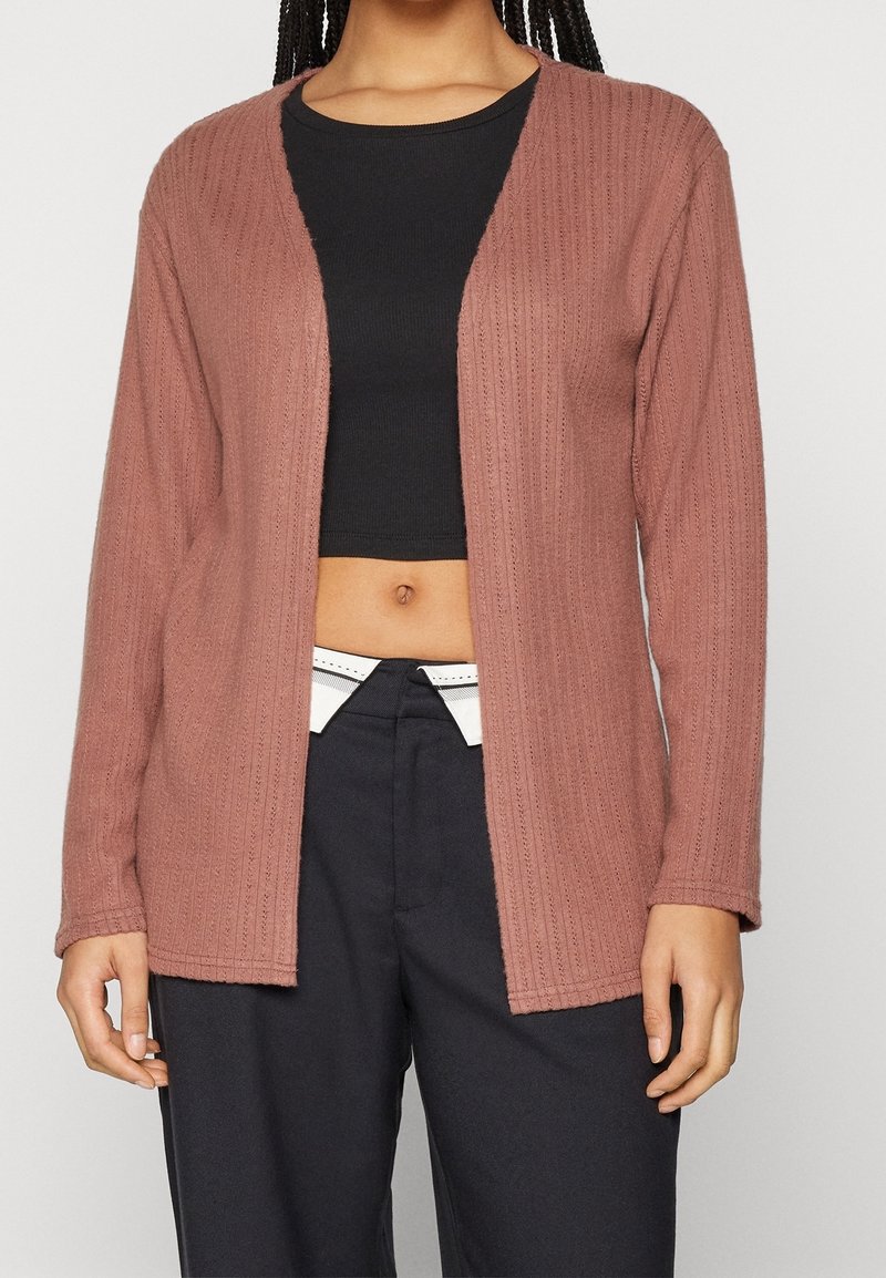 Cardigan en maille couleur rose poudré, avec une texture côtelée, ouverture sur le devant, manches longues et coupe décontractée. Porté avec un crop top noir.