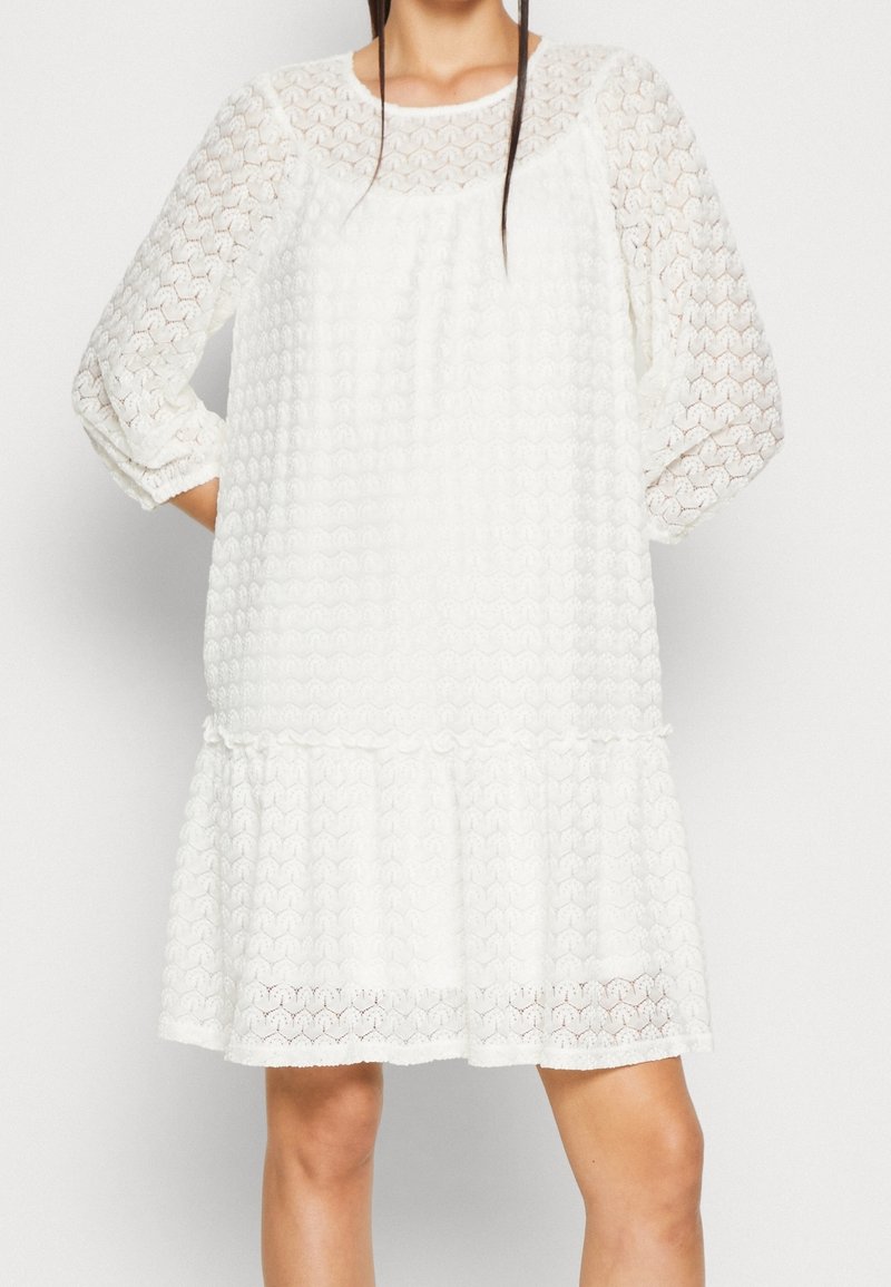 Robe blanche longueur genou avec motif en dentelle hexagonal texturé, manches longues transparentes et coupe ample et fluide sur une femme debout.