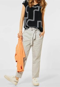 Haut noir à manches courtes avec un motif géométrique blanc, associé à un pantalon gris clair avec taille à lien et poches latérales. Veste orange drapée.