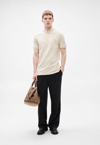 Μπεζ πόλο μπλουζάκι με κοντά μανίκια, χρυσή λεπτομέρεια λογότυπου, συνδυασμένο με φαρδιά μαύρη παντελόνα. Κρατά καφέ δερμάτινη τσάντα με δομημένο σχέδιο.