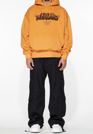 Person mit einem orangefarbenen Kapuzenpullover mit schwarzem Textaufdruck, schwarzen Cargohosen und weißen Sneakers, stehend vor einem weißen Hintergrund.