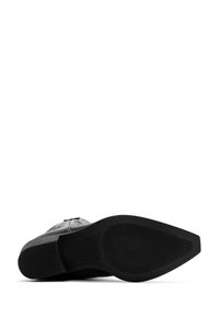 Zapato de cuero negro con una suela texturizada que presenta un patrón estriado, puntera redonda y acentos decorativos en la parte superior.