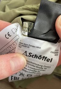 Beige Stoffetikett mit Pflegehinweisen und Branding für ein Hemd, mit schwarzem und grünem Stickmuster. Die Details umfassen Größe und Produktstil.