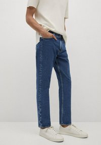 Jeans de mezclilla en un tono azul oscuro, diseño de pierna recta, con bolsillos frontales y costuras sutiles, combinados con zapatillas blancas y una blusa clara.
