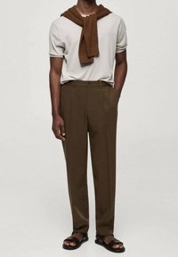 Kurzärmliges, hellgraues Hemd mit einem braunen, gestrickten Pullover, der über die Schultern gelegt ist. Braune, maßgeschneiderte Hose und schwarze Sandalen vervollständigen das Outfit.