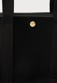 Černá textilní taška s širokými strukturovanými popruhy, podrobným šitím a malým kulatým zlatým knoflíkem s rytým textem.