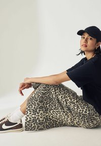 Schwarzes Kurzarmhemd, midi Rock im Leopardenmuster in Beige und Braun, kombiniert mit weißen Socken und dunklen Turnschuhen. Lässiges Outfit, sitzende Pose.