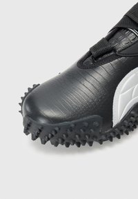 Gros plan d'une chaussure noire texturée avec un panneau latéral blanc et une semelle équipée de grosses pointes en caoutchouc pour l'adhérence.