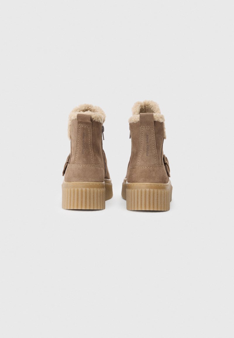 Botas de tobillo de ante marrón con forro de piel de oveja, suelas de goma acanaladas y cremalleras laterales. La vista trasera muestra suave pelaje en el cuello.