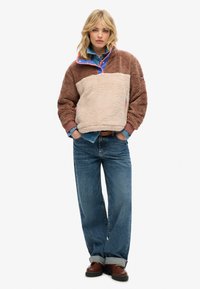 Pull en polaire brun et beige avec un col zippé, un accent rose et une doublure bleue ; porté sur une chemise en denim et assorti à un jean en denim large.