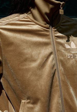 Gros plan d'une veste de survêtement Adidas en velours marron avec fermeture éclair, col montant et logo brodé sur la poitrine.