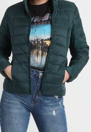 Person iført grøn quiltet jakke over t-shirt med grafisk bylandskab og blå slidte jeans, med hænderne i jakkelommerne.