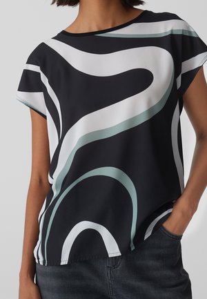 Woman wearing a black top with white and light green abstract curved stripes, paired with dark denim jeans, hand in pocket.